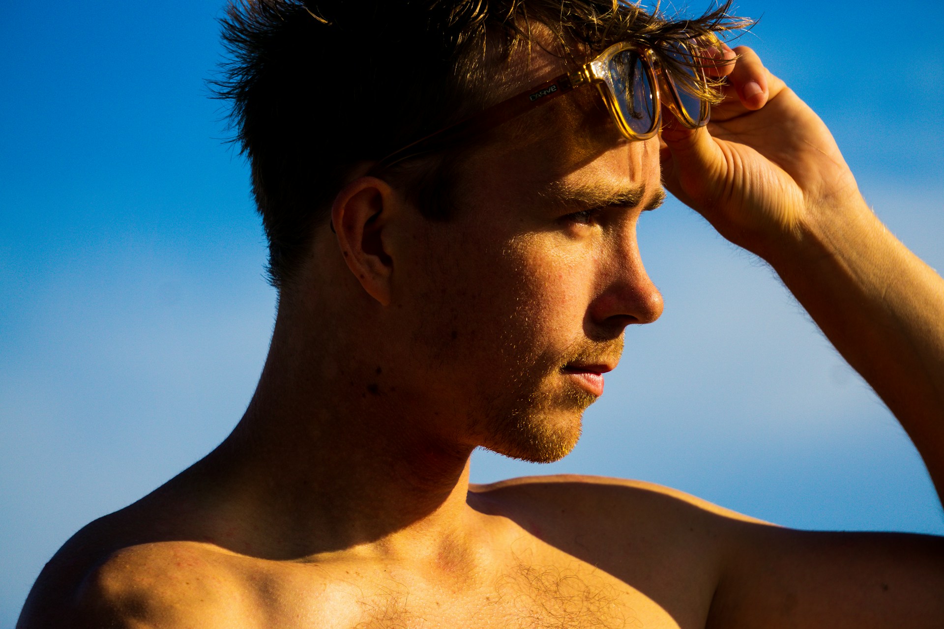 Homme de profil sous un ciel bleu, main sur le front et lunettes de soleil relevées, peau exposée au soleil d'été